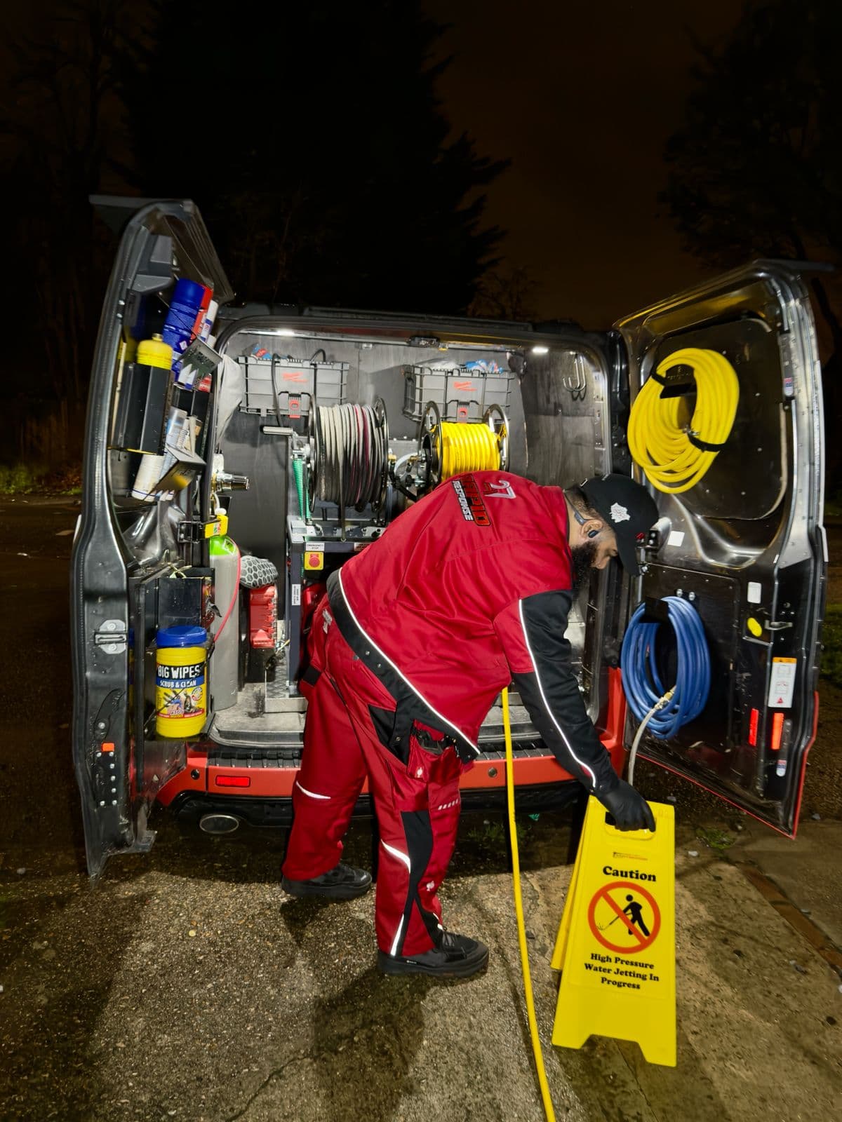 247 Rapid Response engineer at the rear of a fully-stocked branded van, drainage jetting equipment loaded and ready, on a London street at night